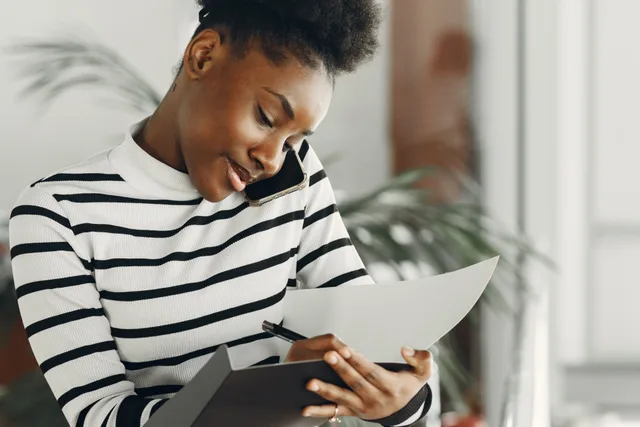 Young African woman multitasking with a phone call and taking notes indoors, wearing stripes. 3 parts of local delivery discovery call, explained