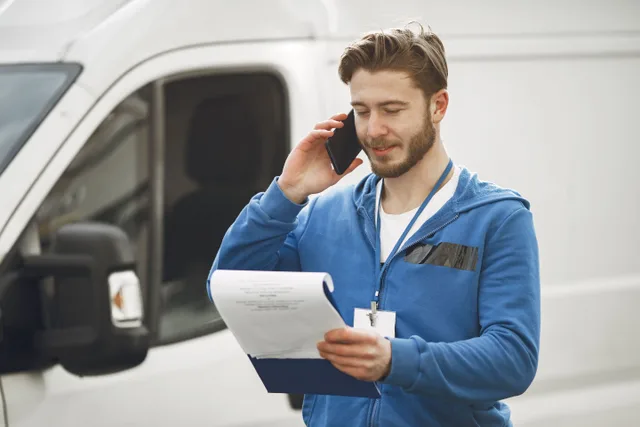 A delivery man in a blue hoodie makes a phone call while holding a clipboard, standing by a white van. Starting a local delivery business: choosing a model, insurance, and permits