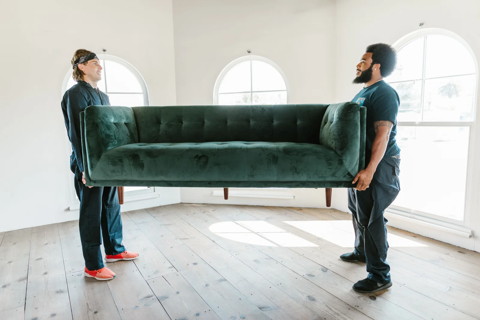 Two men lifting a green sofa in a well-lit room with arched windows. White glove delivery pricing per stop in the US: ranges and add-ons